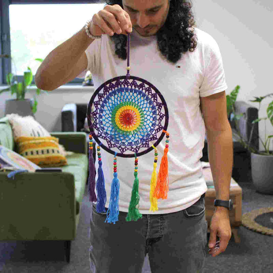 Person holding a small, vivid rainbow dreamcatcher in a room with plants and furniture.