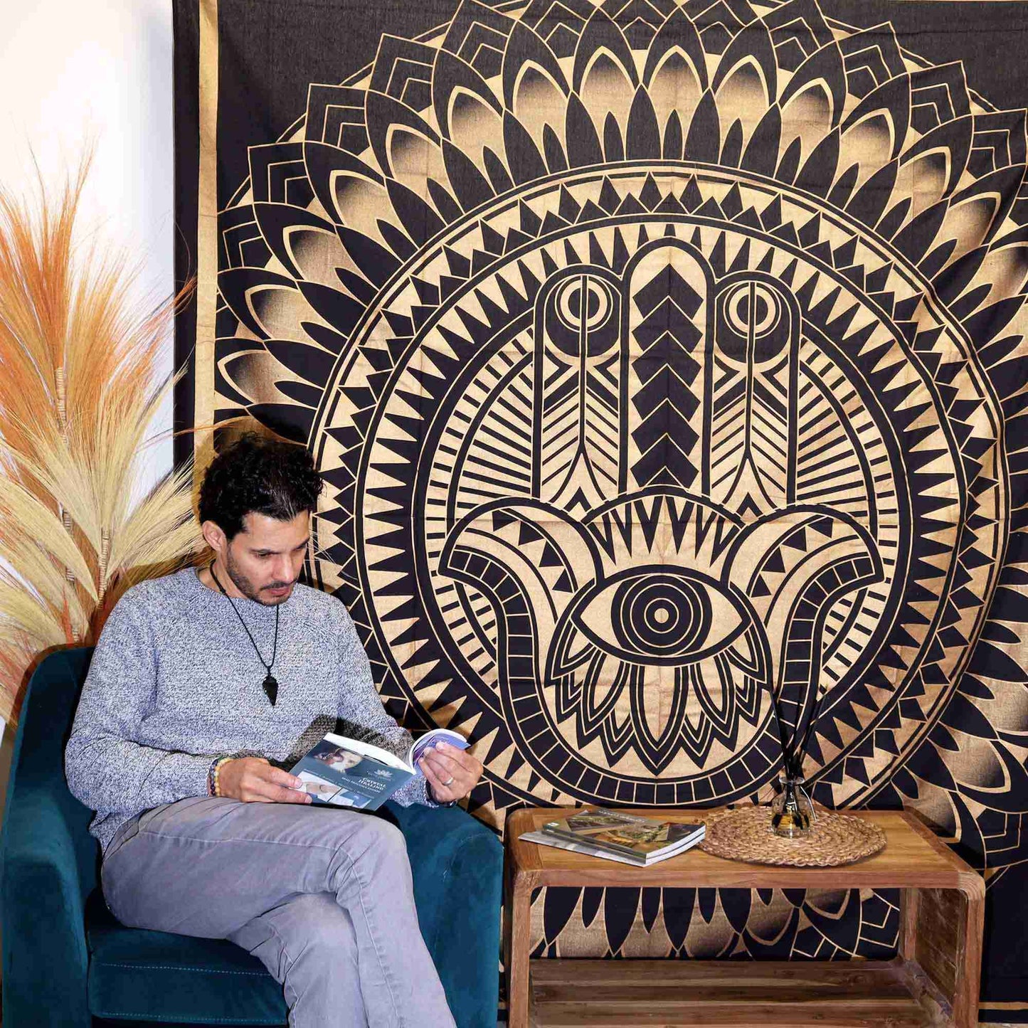 Man sitting on a blue chair reading a book in front of a Double Cotton Bedspread + Wall Hanging - Hamsa - Black Gold