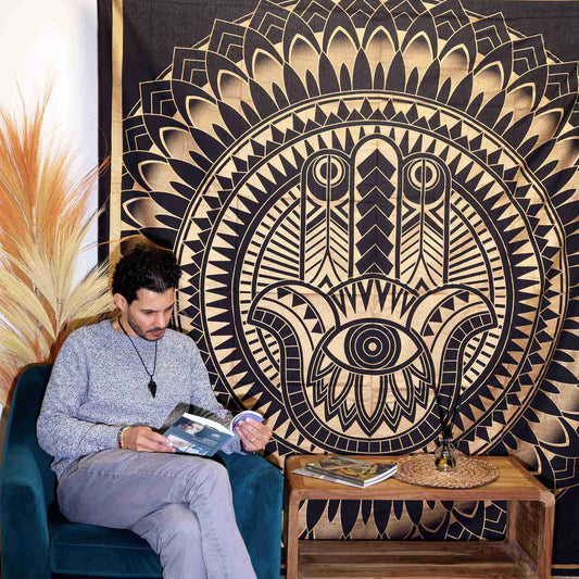 Man sitting on a blue chair reading a book in front of a Double Cotton Bedspread + Wall Hanging - Hamsa - Black Gold