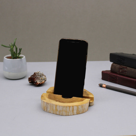 Wooden phone stand holding a smartphone on a desk with a plant and books in the background.