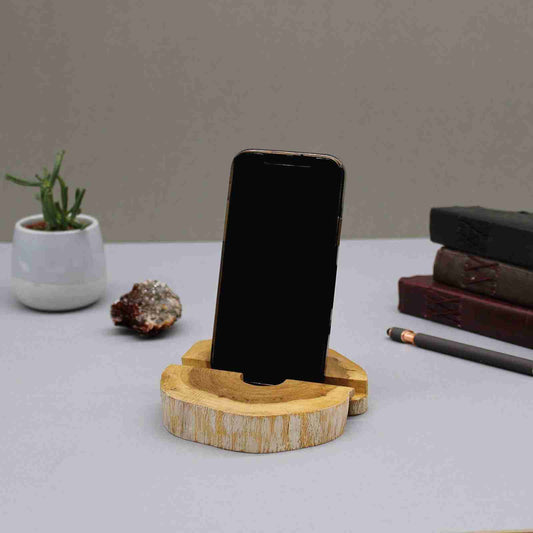 Wooden phone stand holding a smartphone on a desk with a plant and books in the background.