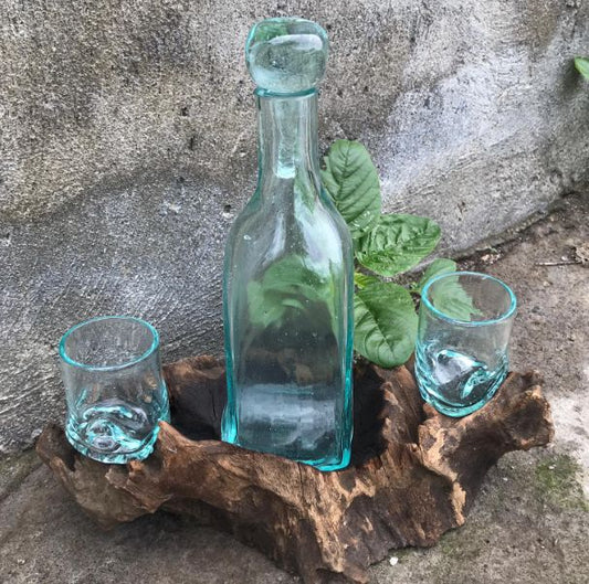 One Turquoise glass bottle and two glasses on a piece of gamal wood with greenery against a stone wall.