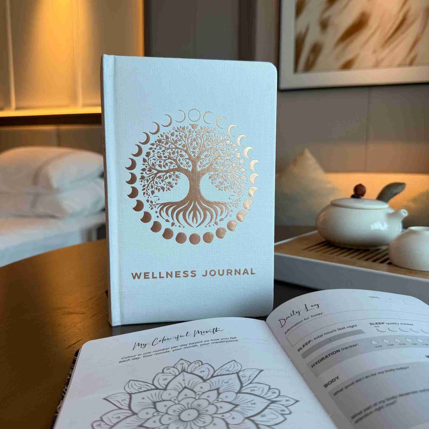 Wellness journal with tree of life design on cover, placed on a table with a tea set in the background.