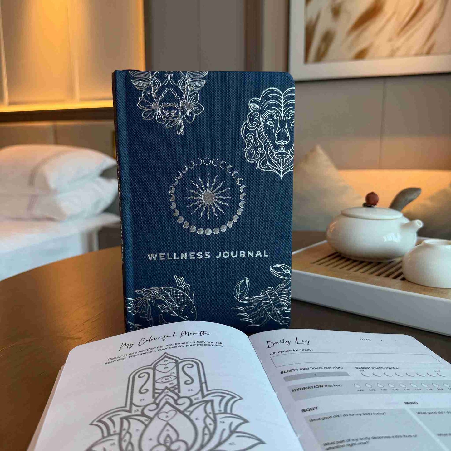 Blue wellness journal with white designs on a table with a teapot and cups in the background.
