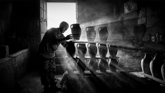 Artisan arranging handmade clay pots in a pottery workshop, with sunlight streaming through the doorway and illuminating the dust in the air.