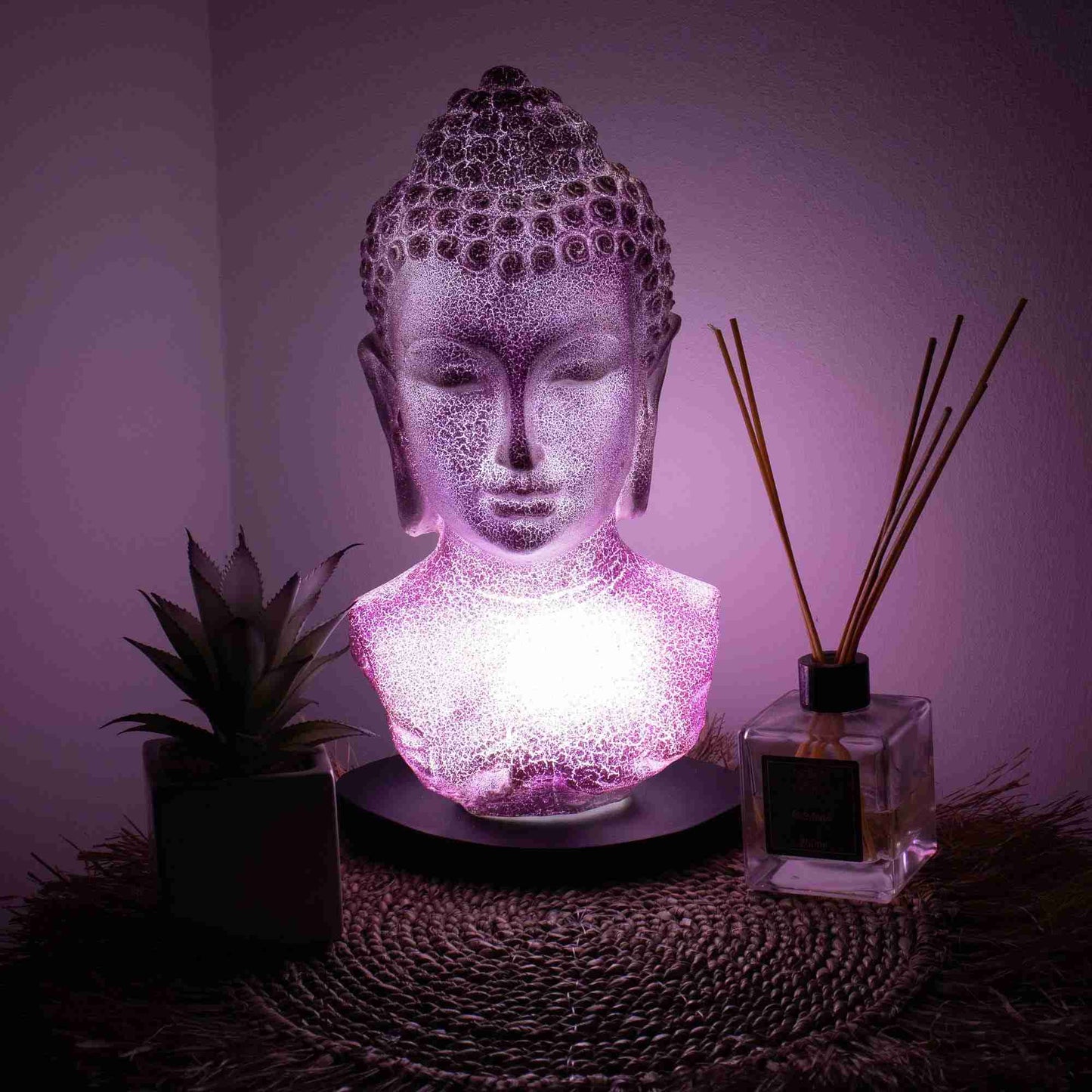 Buddha Bust Lamp, Lavender Crackle with light on, with a plant and diffuser on a textured surface.