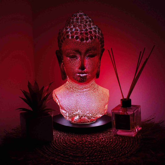 Decorative lamp shaped like a Buddha bust with a red glow, placed on a surface with a plant and diffuser in the background.