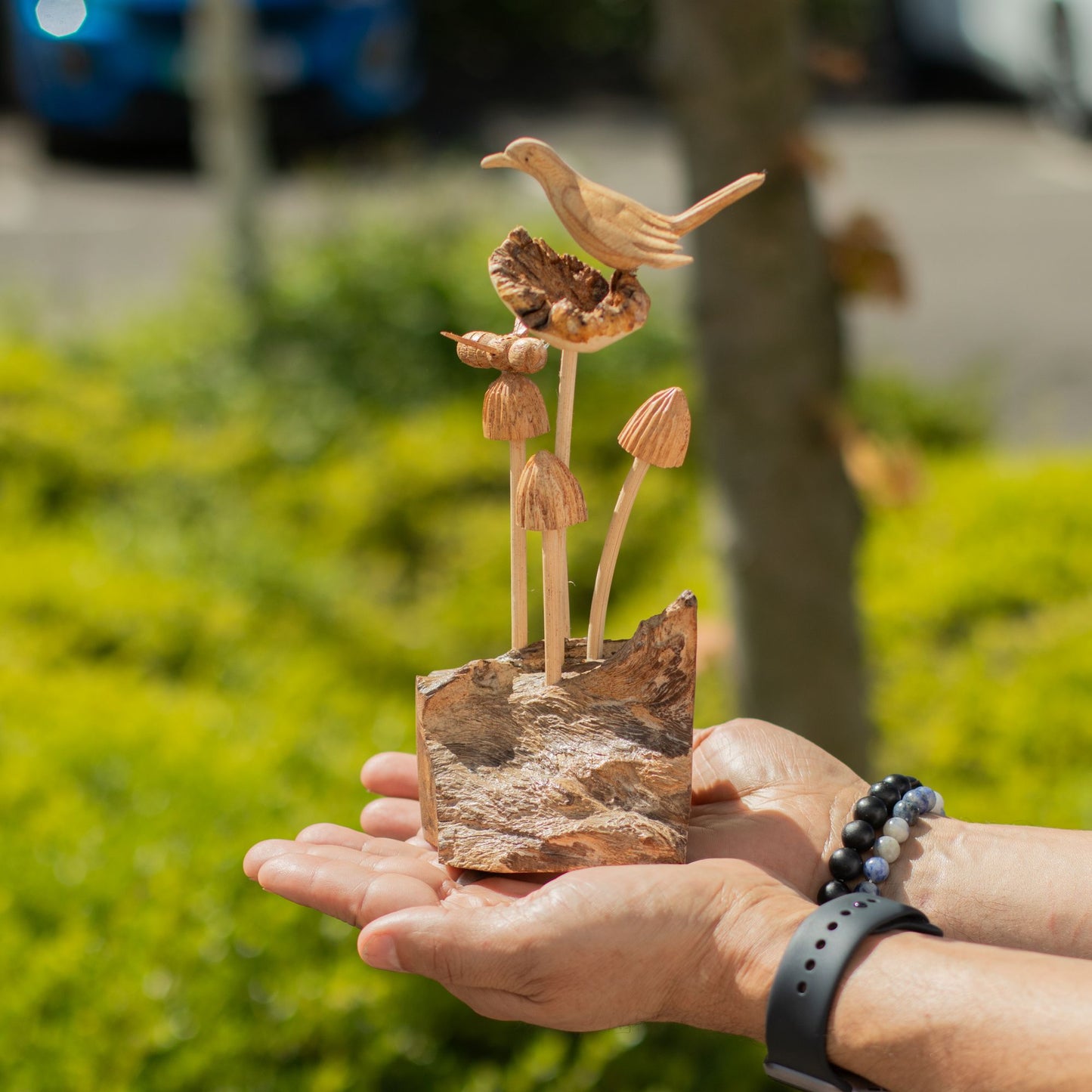 Handmade Parasite Wood Art Carving Bird & Bee on Mushrooms held in hands with a blurred natural background