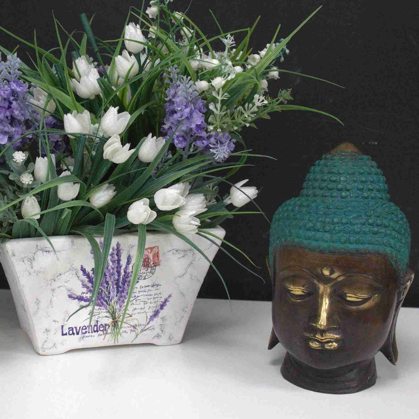 Large Brass Buddha Head Figurine Classic next to a potted plant with white and purple flowers on a white surface.
