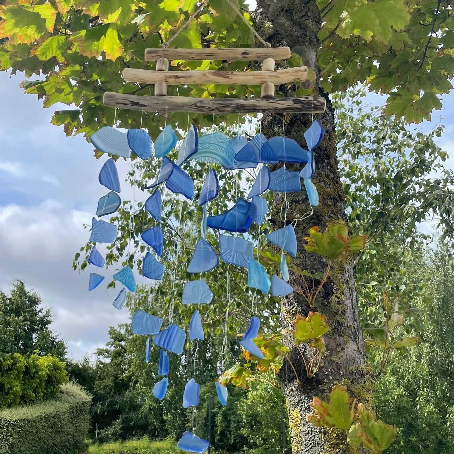 Three Stick - All Blues Glass Wind Chime hanging from a tree with green leaves and sky in the background