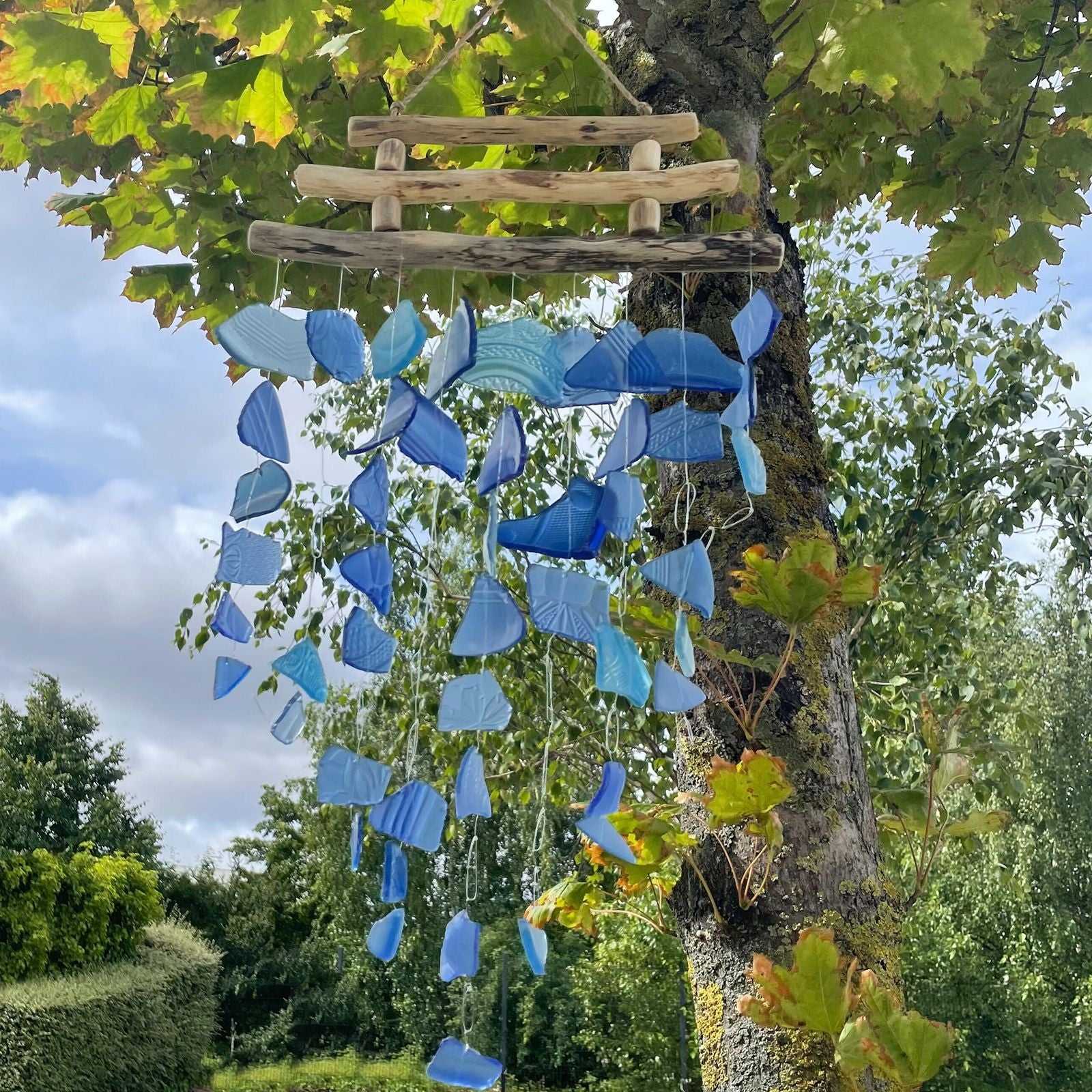 Three Stick - All Blues Glass Wind Chime hanging from a tree with green leaves and sky in the background