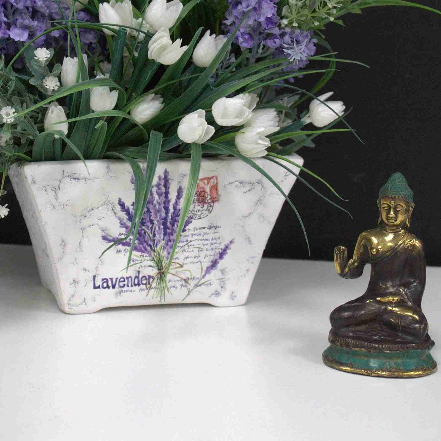 Decorative planter with lavender and tulips next to a Mini Hand Up Sitting Buddha Brass Figurine on a white surface.