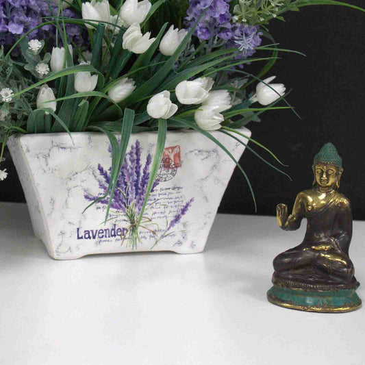 Decorative planter with lavender and tulips next to a Mini Hand Up Sitting Buddha Brass Figurine on a white surface.
