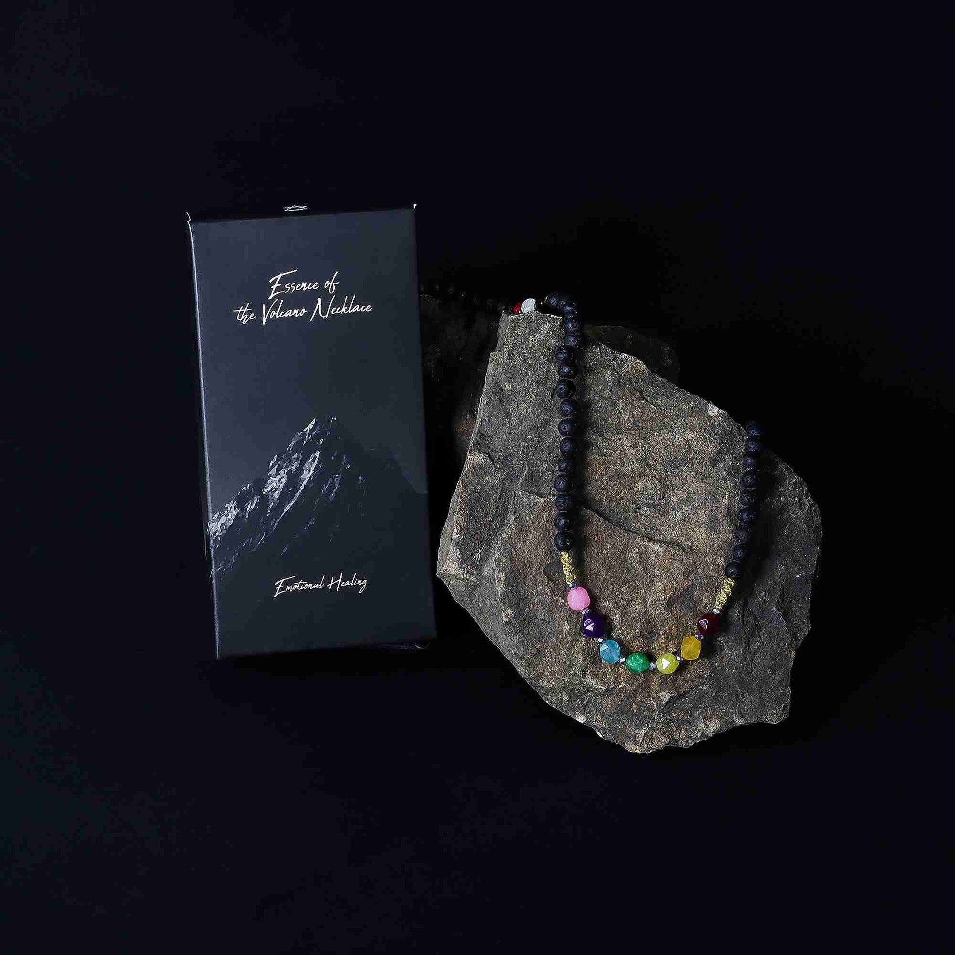 Necklace with colorful beads on a rock next to a box labeled 'Exodus of the Volcano Necklace' on a black background