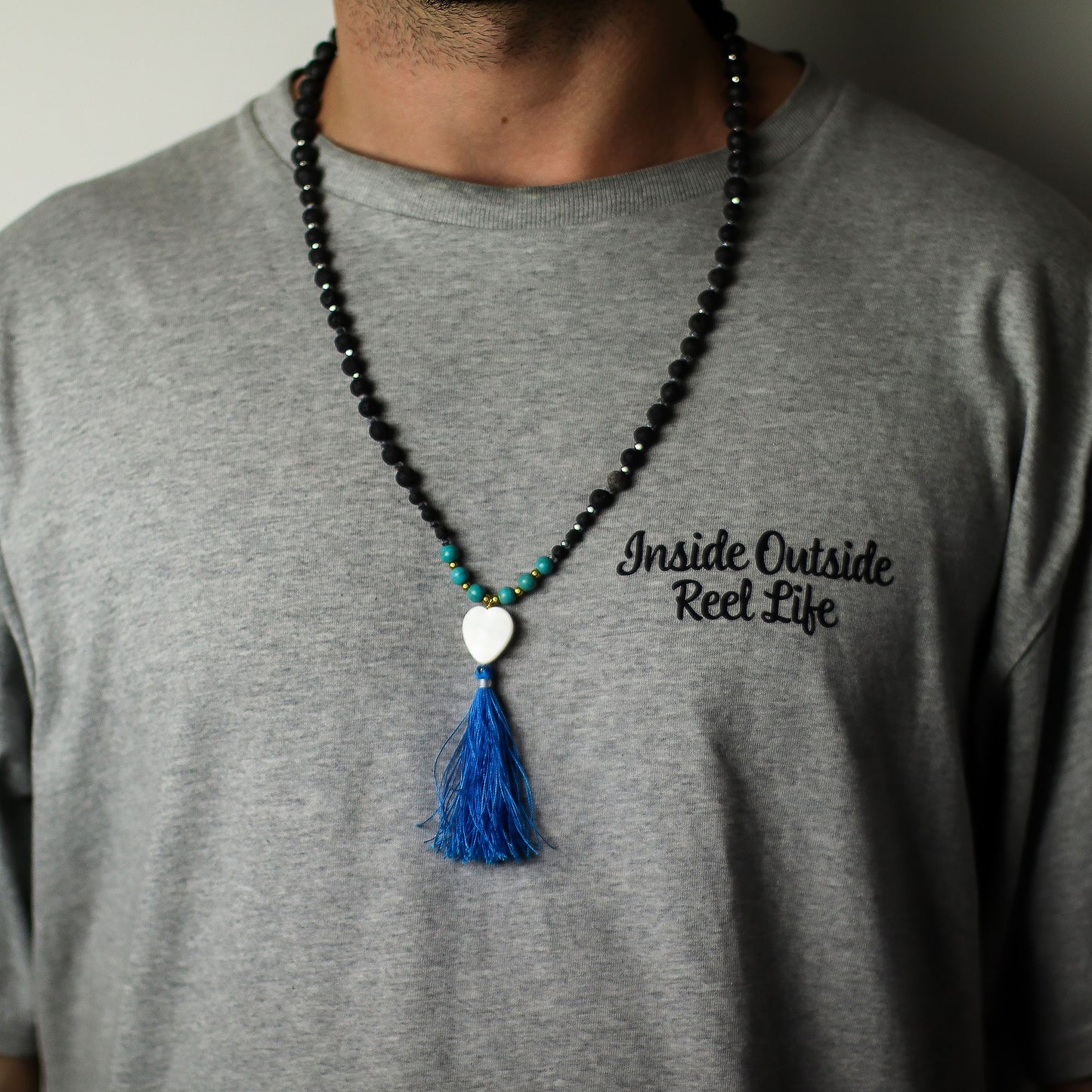 Person wearing a gray t-shirt with text and a beaded essence of volcano necklace with a blue tassel.