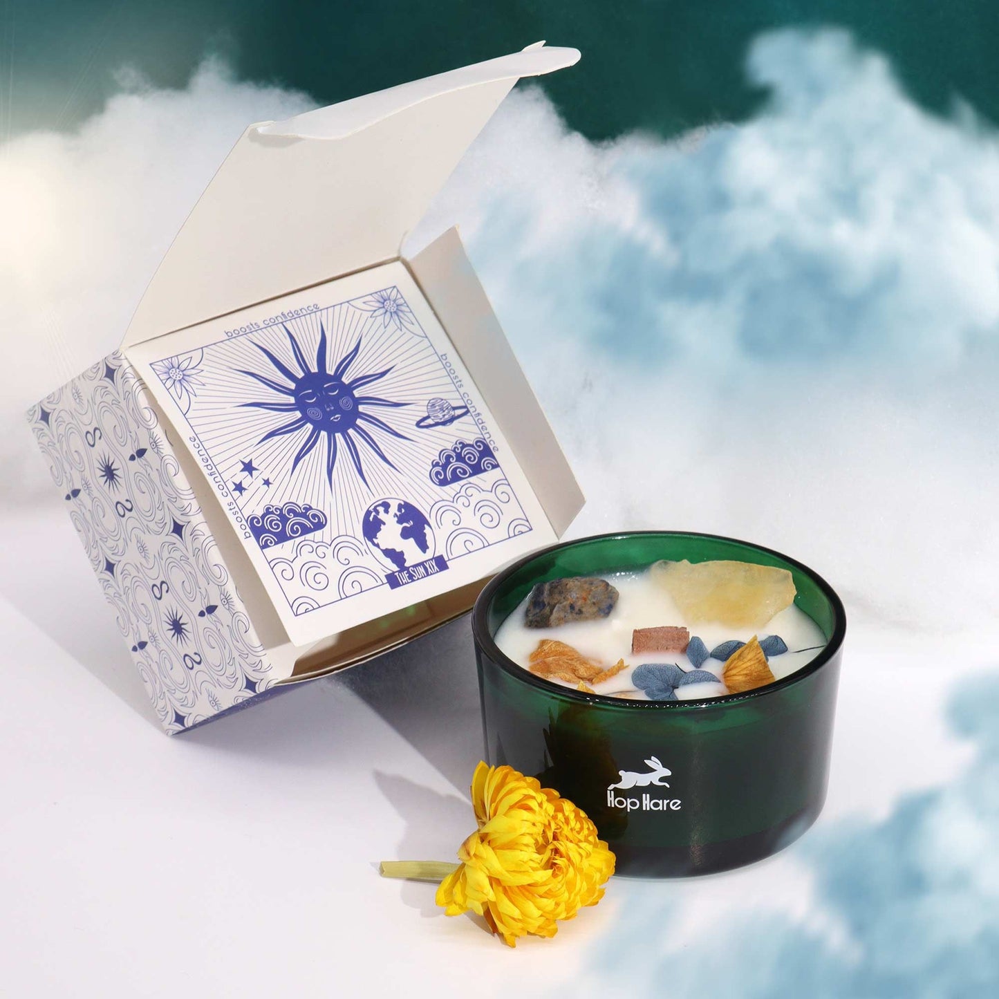 Hop Hare Crystal Magic Flower Candle - The Sun, next to an open box with a sun design, on a cloud background.