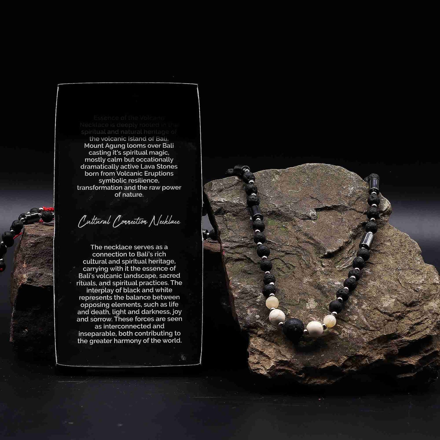 Essence Of The Volcano Necklace with black and white beads on a rock next to an information card