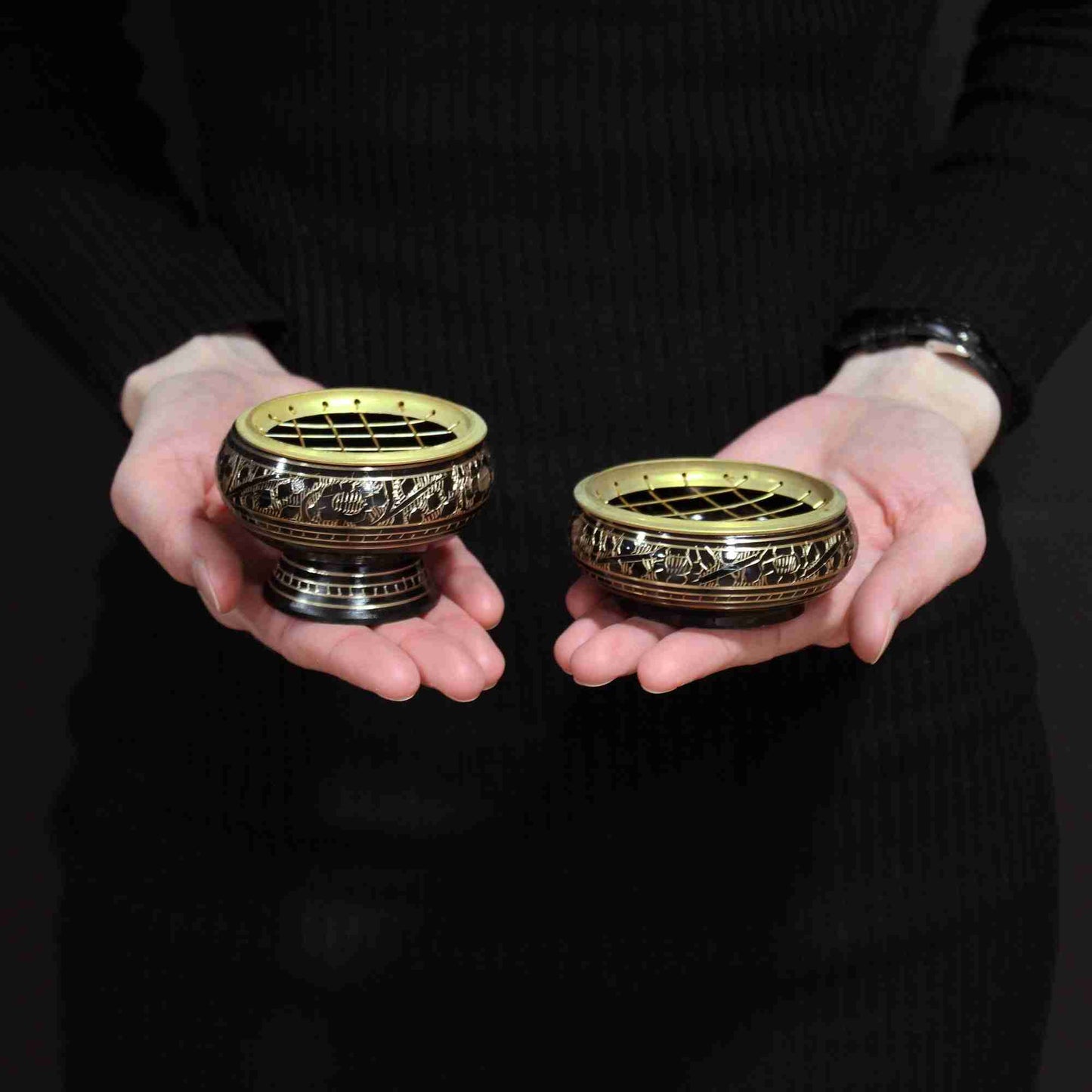 Two brass incense burners held in hands against a dark background