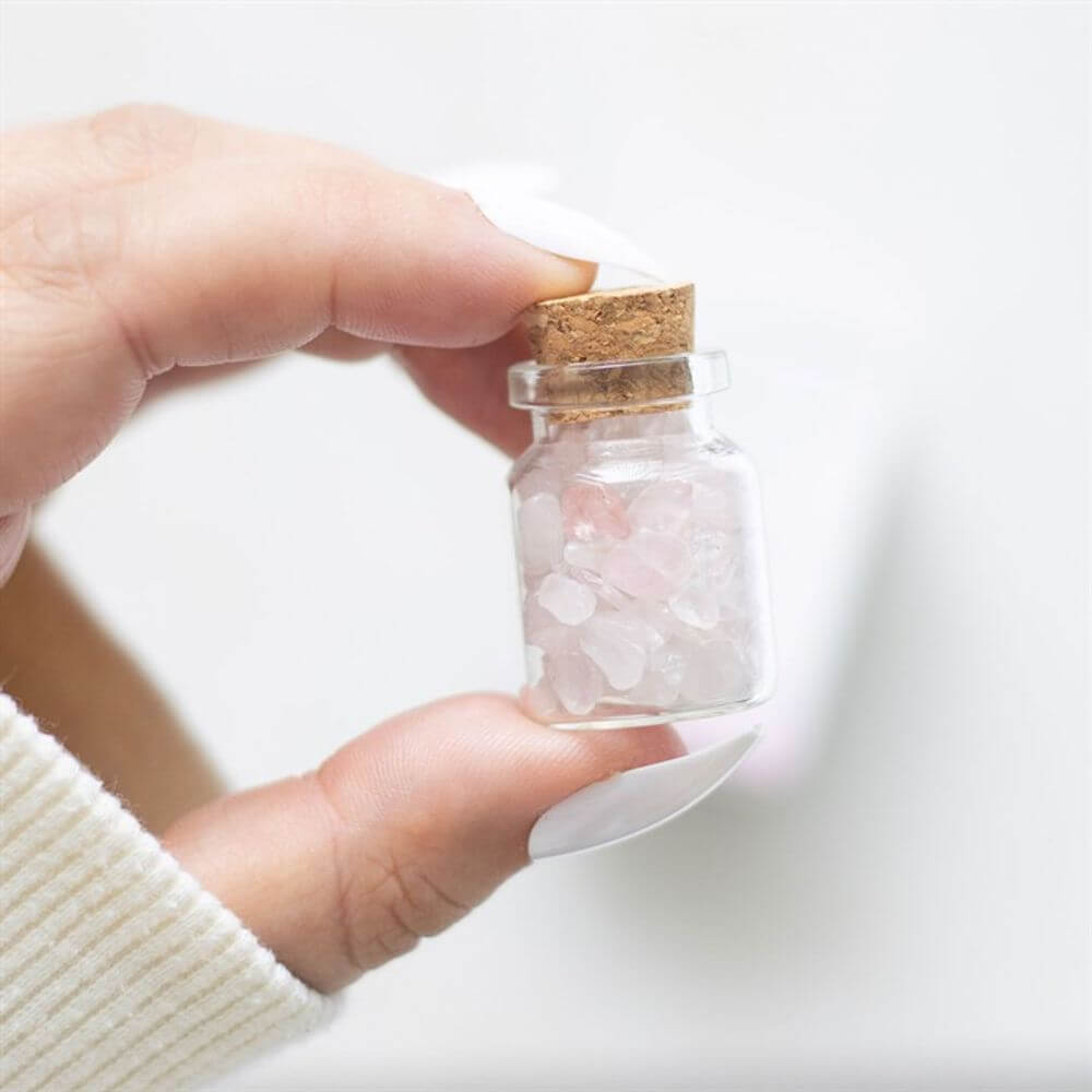 Hand holding a small jar of rose quartz crystal chips with a cork lid, promoting love and healthy relationships.